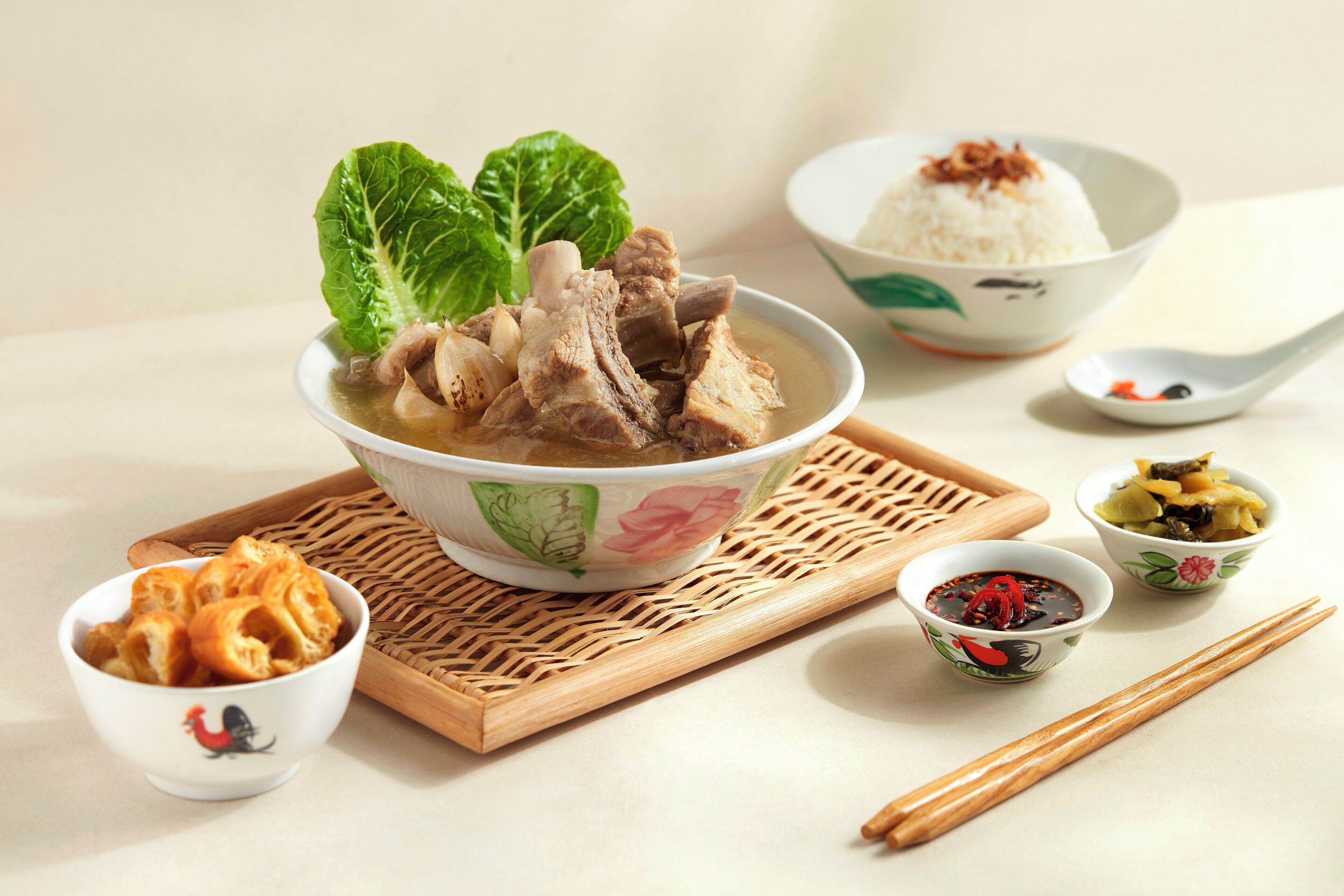 A bowl of soup with meat and vegetables, a bowl of rice, and two small bowls of condiments on a woven mat.