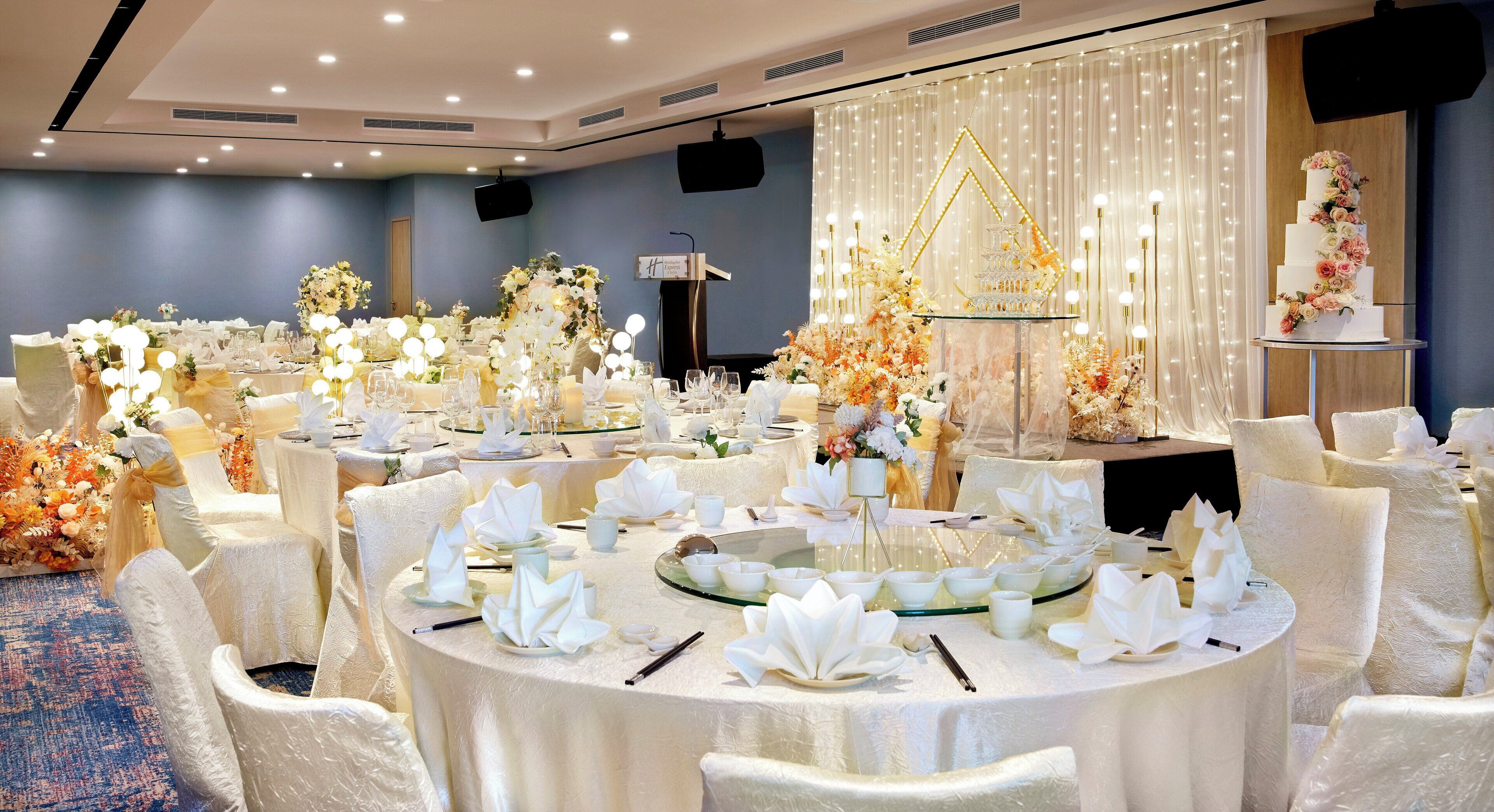 A banquet hall with round tables set for a formal event, featuring white tablecloths, floral centerpieces, and a tiered cake on a stand.