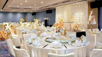 A banquet hall with round tables set for a formal event, featuring white tablecloths, floral centerpieces, and a tiered cake on a stand.