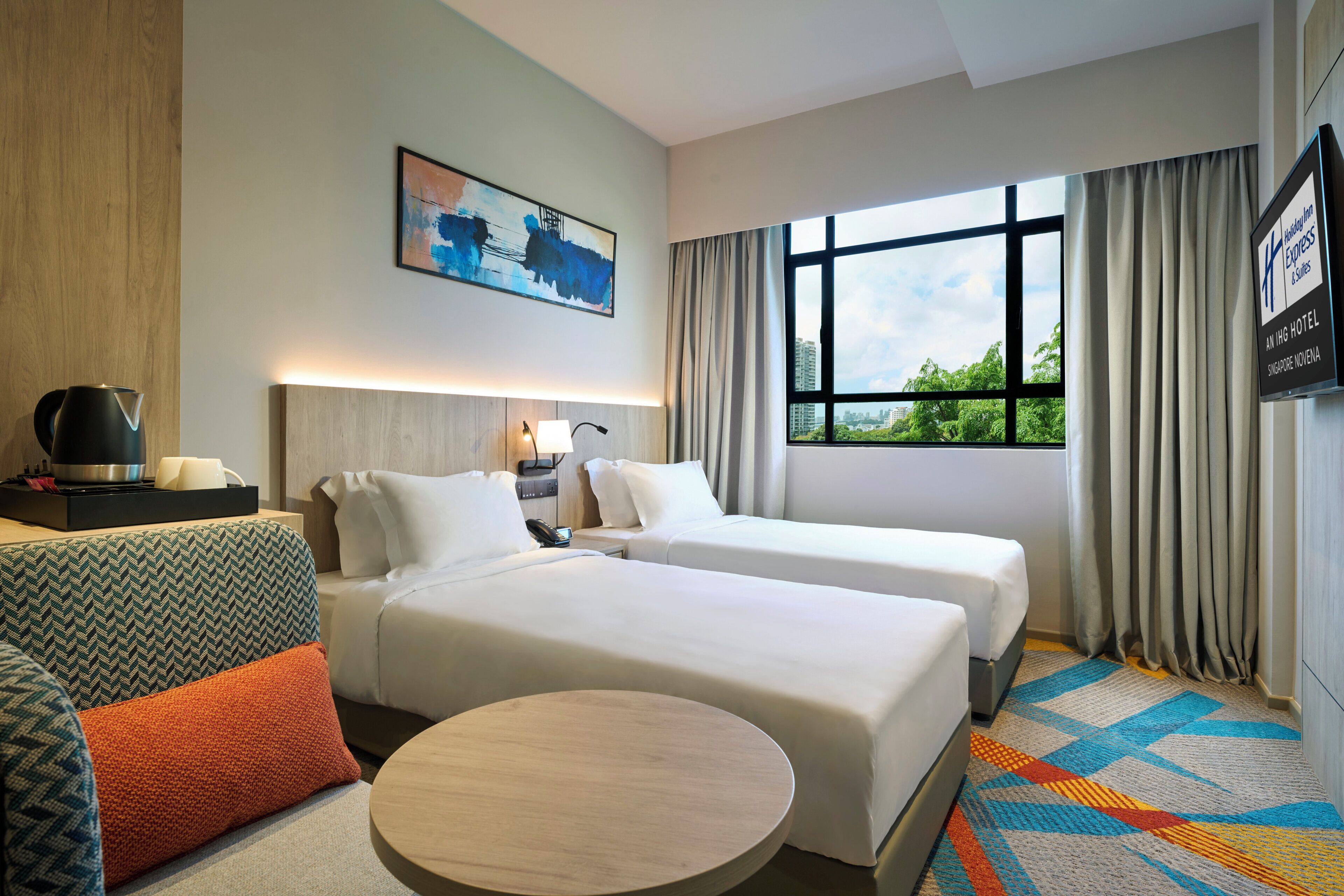 Une chambre d’hôtel avec deux lits, un canapé, une table ronde, un tableau au mur et une vue sur la verdure par la fenêtre.
