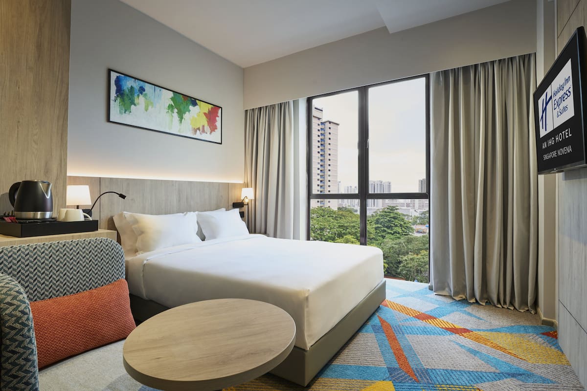 Hotelkamer met een bed, een rond tafel, een bank, een koffiezetapparaat en uitzicht op de stad door grote ramen.