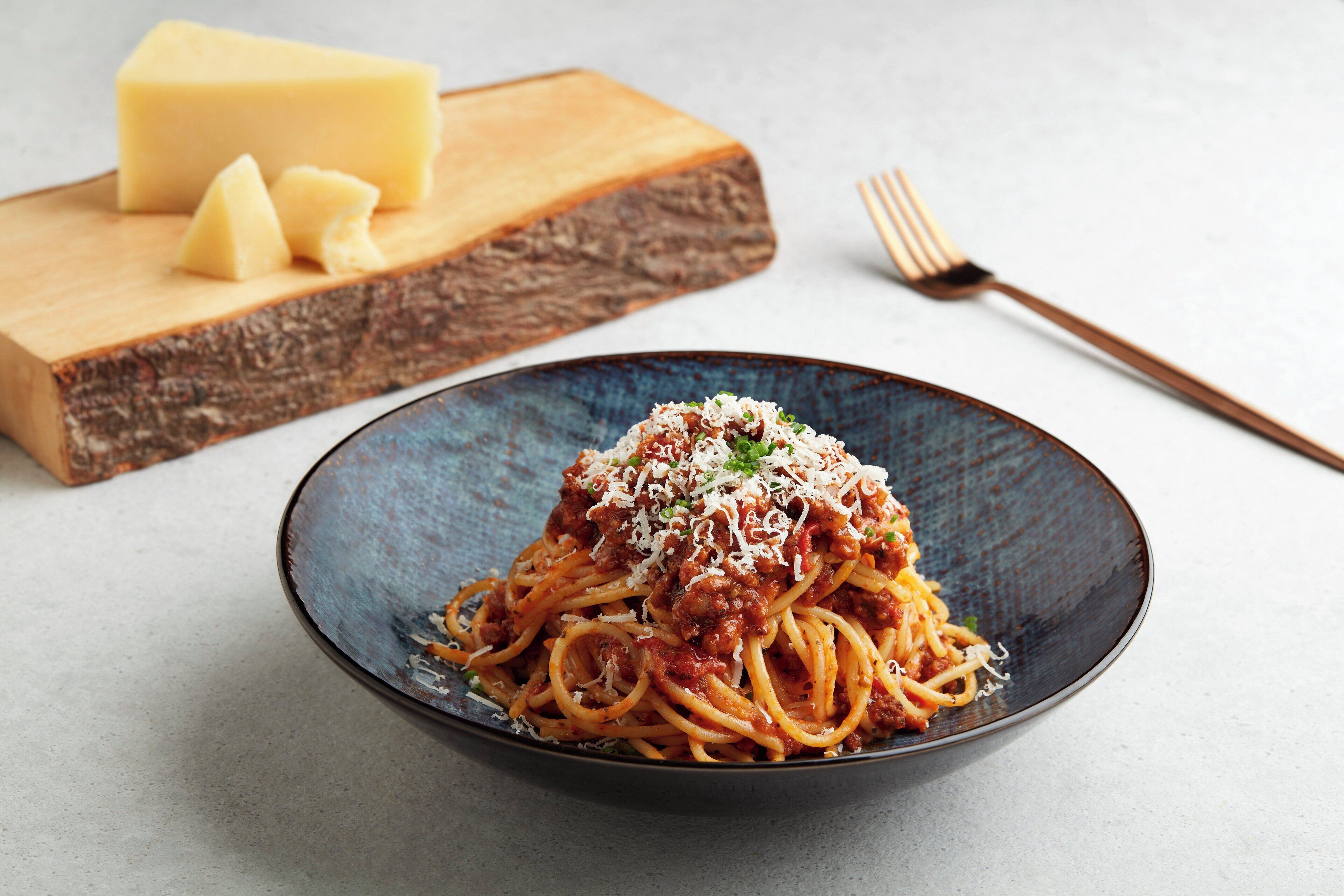 Een bord spaghetti met vleessaus en geraspte kaas, een houten snijplank met een blok kaas en kaasvlokken, en een vork op een tafel.