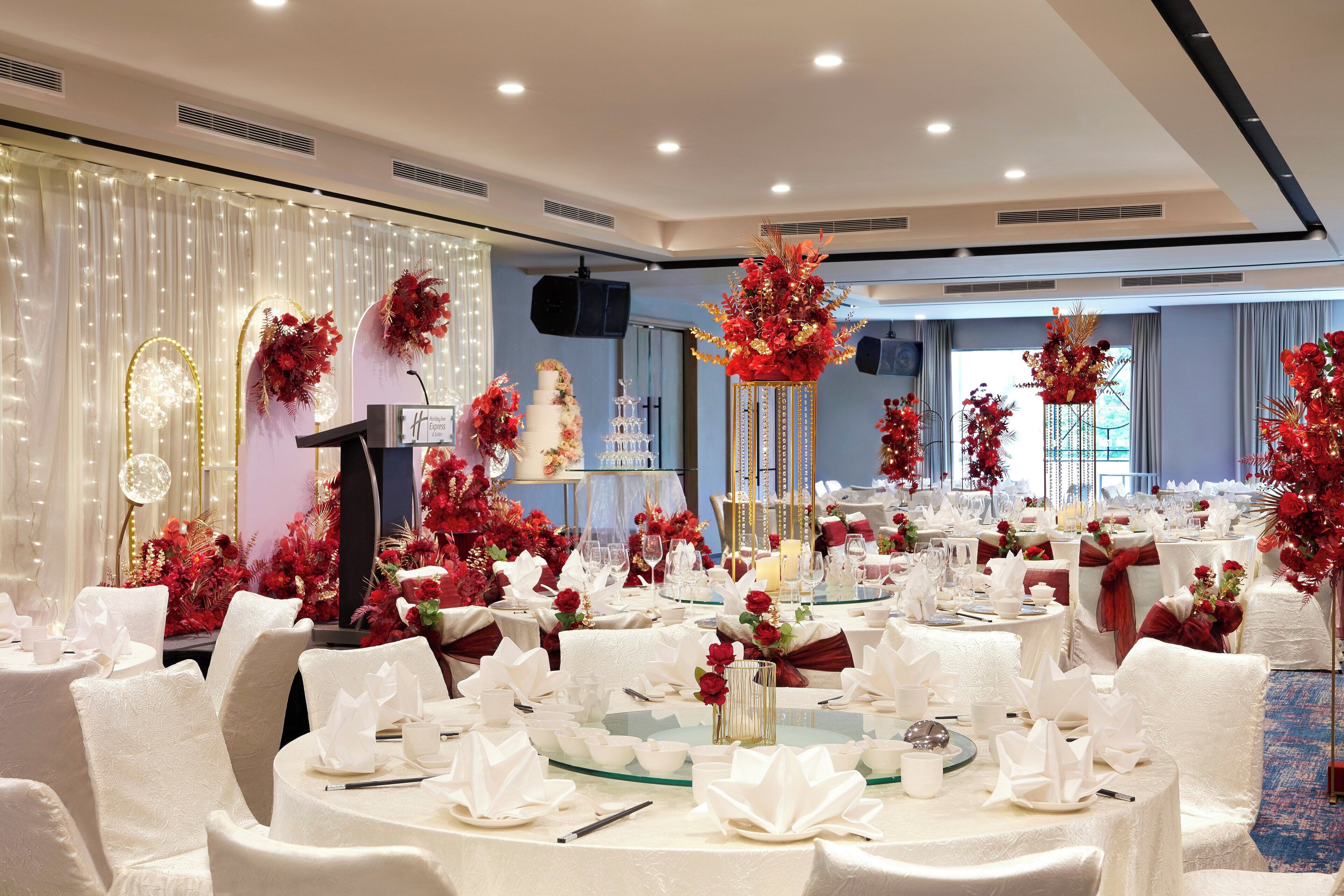 A banquet hall with round tables, white tablecloths, and red floral centerpieces.