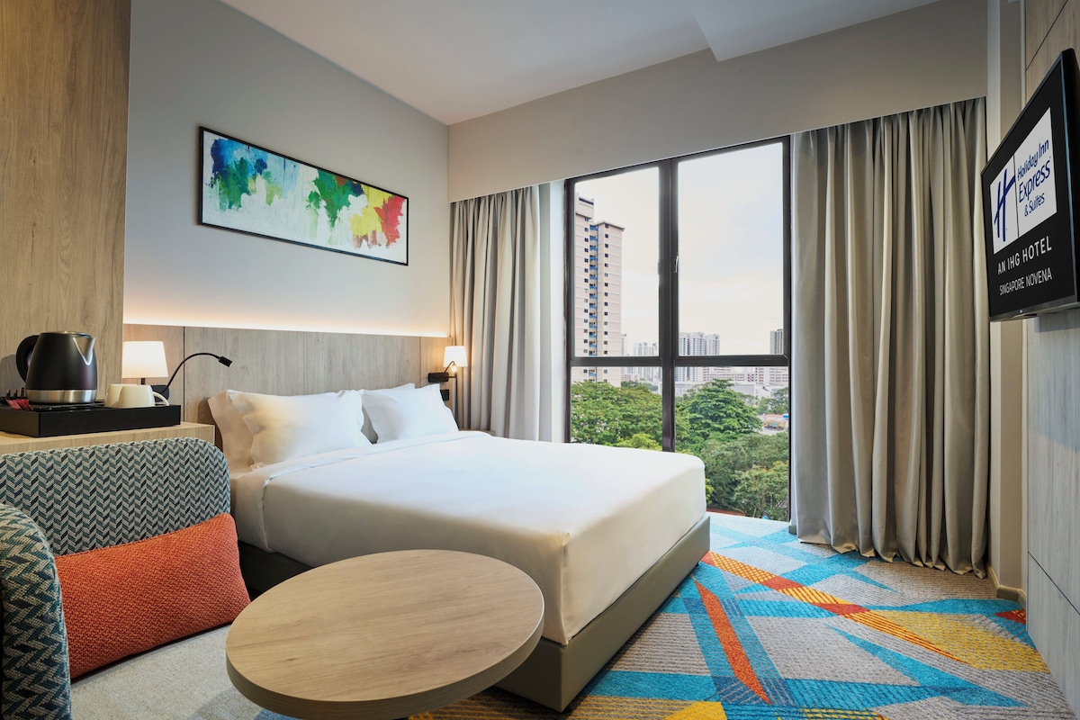 Ein Hotelzimmer mit Bett, rundem Tisch, einer Couch, einer Kaffeemaschine und Blick auf die Stadt durch große Fenster.