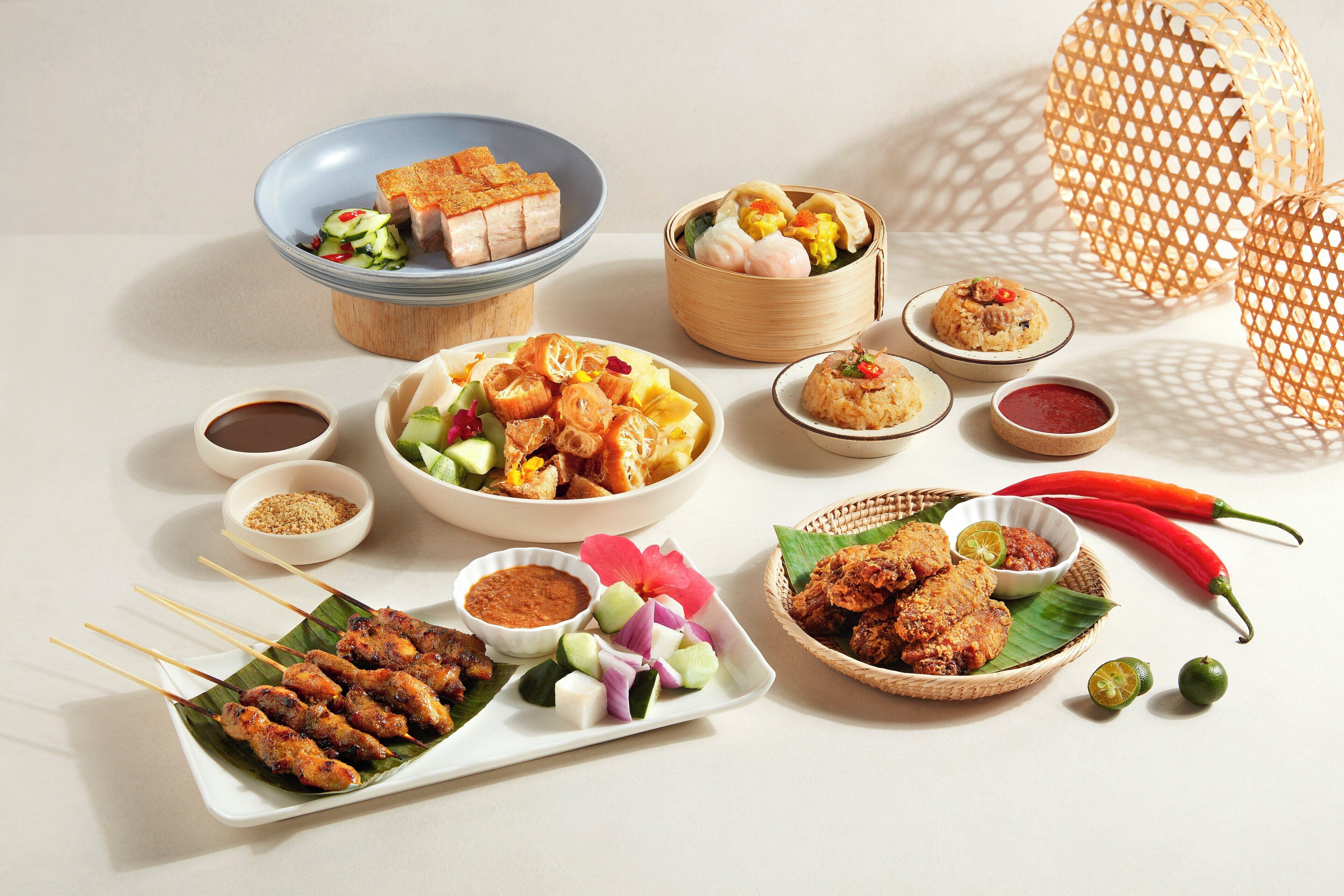 A variety of dishes including skewers, a bowl of soup, and a plate of fried items, arranged on a table with a woven basket and a red chili pepper.
