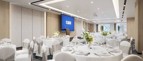 A conference room with multiple round tables set for dining, white chairs, and a large screen displaying a presentation.