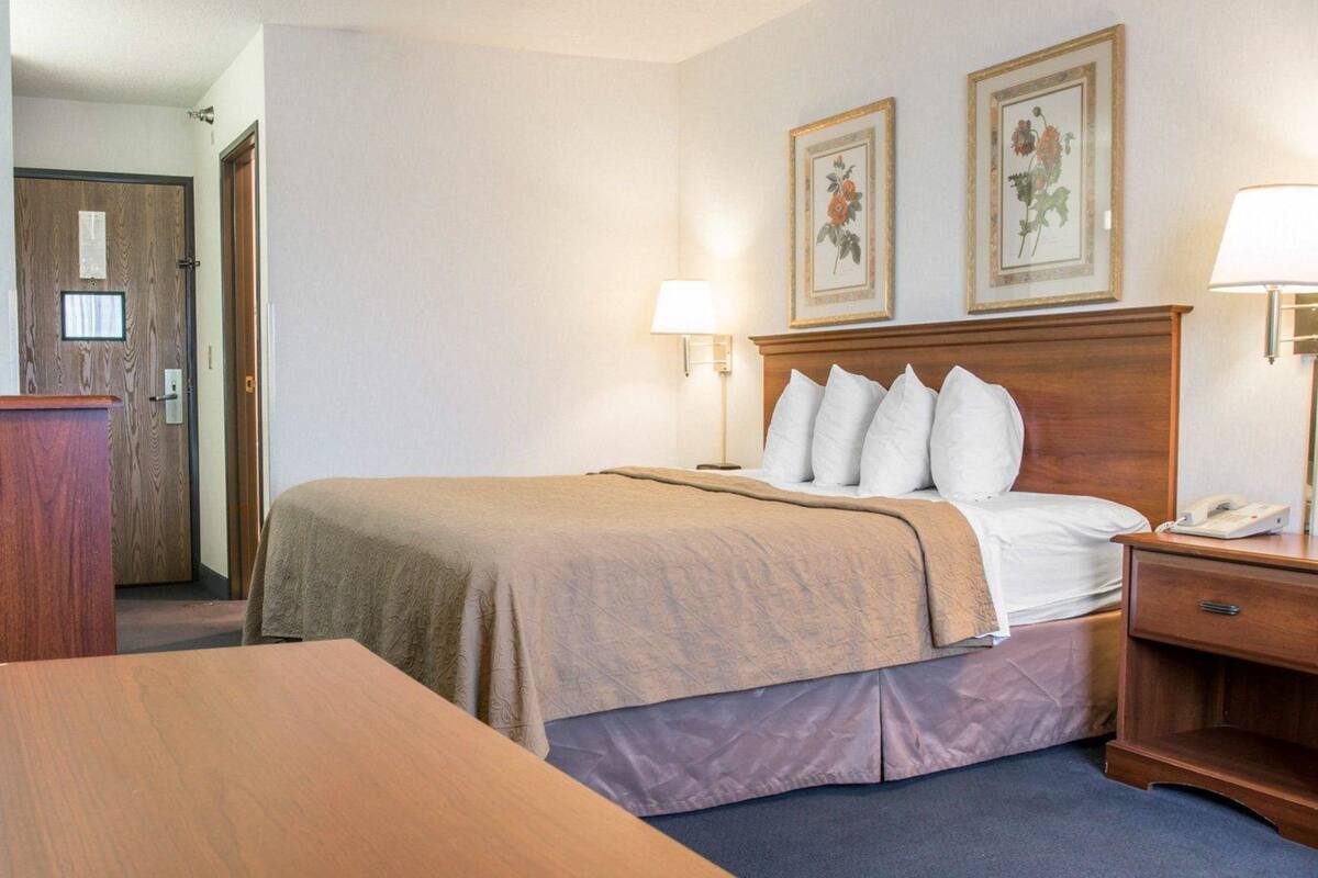 A hotel room with a bed, bedside lamps, a wooden headboard, a desk, and a door.