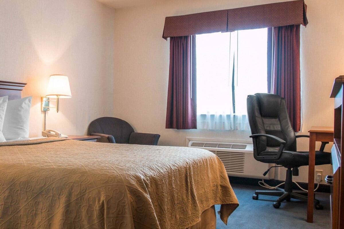 A hotel room with a bed, a desk, a chair, a lamp, and an air conditioning unit.