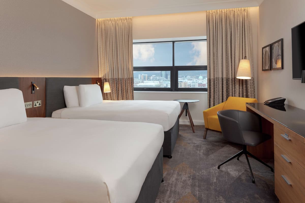 Habitación de hotel con dos camas, un escritorio, una silla, un televisor y una ventana con vista a la ciudad.