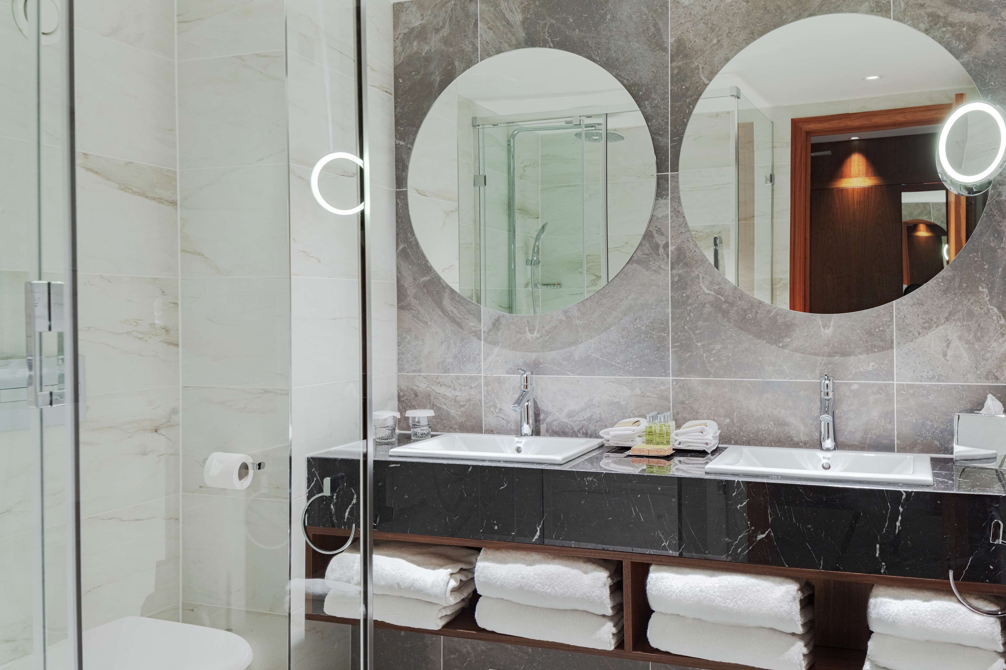 Un baño moderno con ducha de vidrio, tocador con dos lavabos y un espejo grande con marcos circulares.