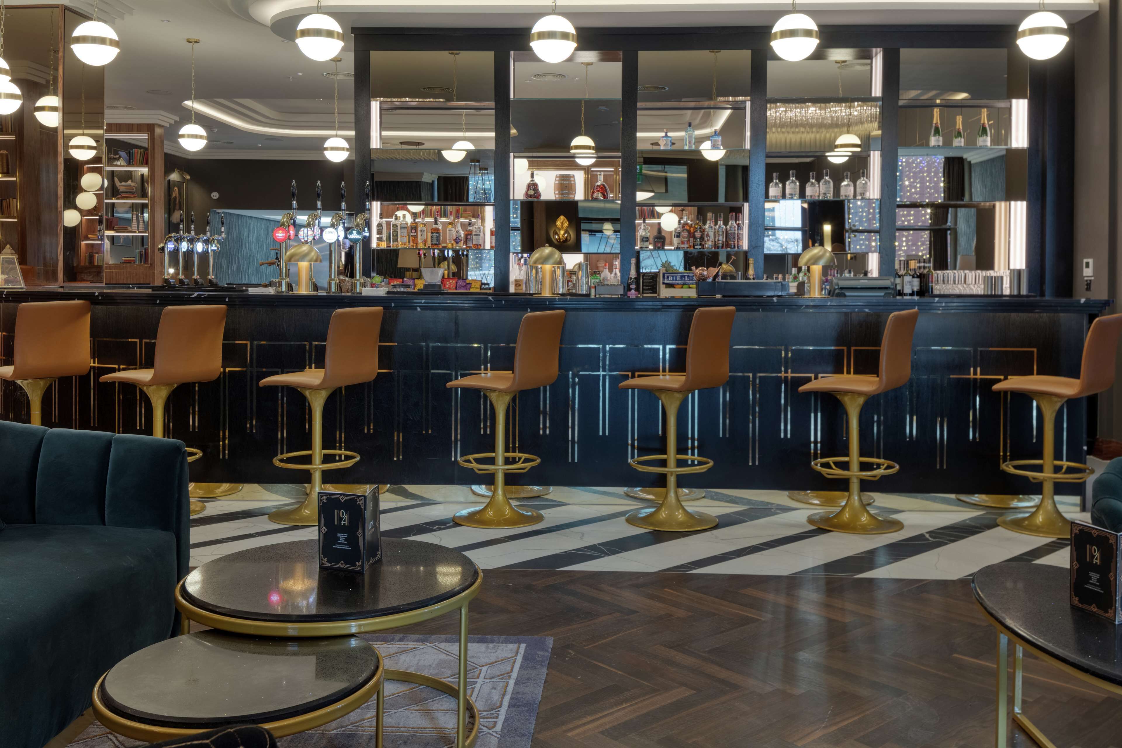 Un espace bar doté de plusieurs tabourets, d’un comptoir avec des bouteilles et d’un coin salon avec un canapé et des tables.