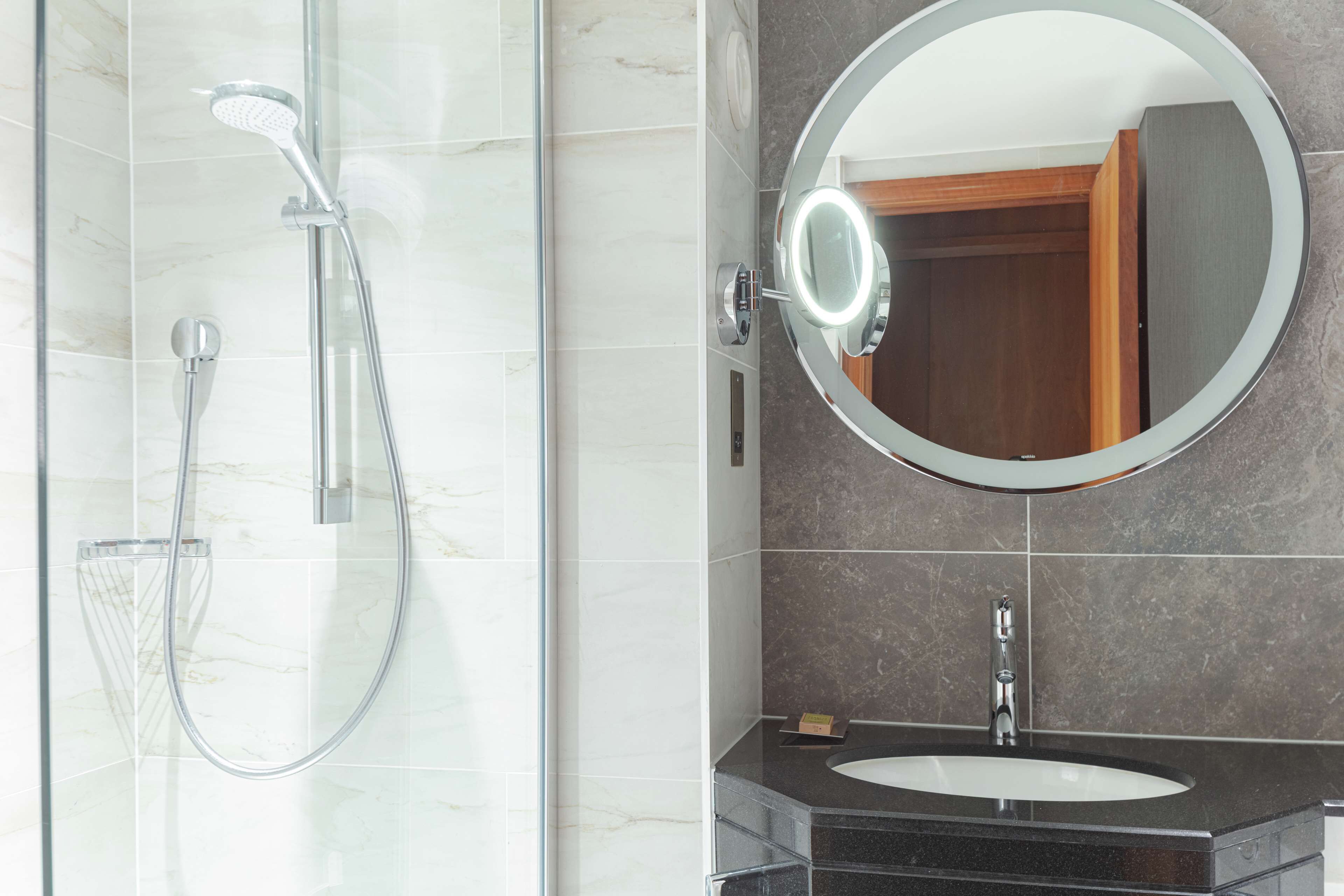 Un baño moderno con ducha envuelta en vidrio, espejo redondo con luz y lavamanos sobre encimera oscura.