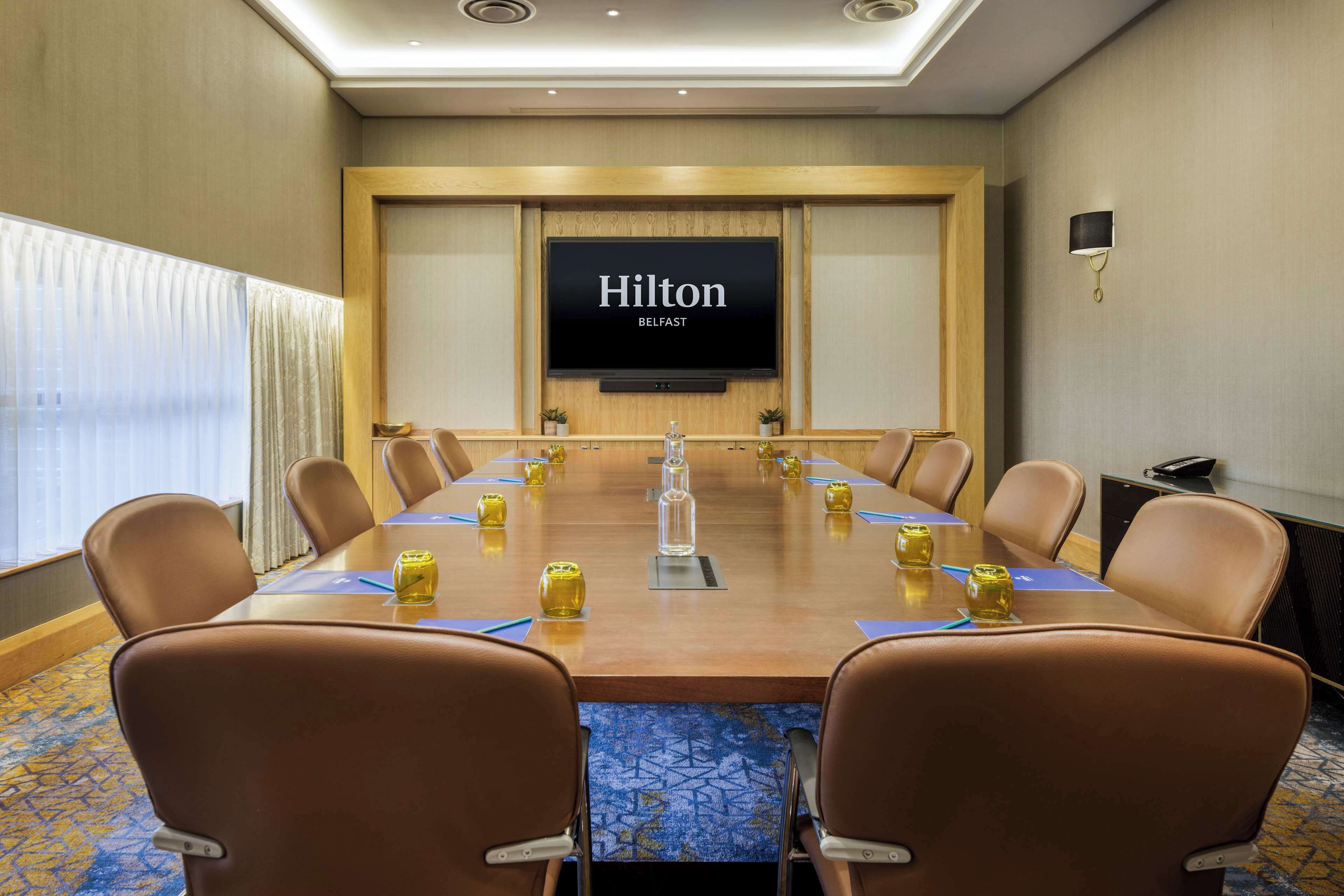 A conference room with a long table, brown leather chairs, a large screen displaying "Hilton Belfast," and a small potted plant on the table.