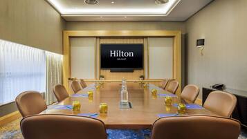 Un salón de conferencias con una larga mesa, sillas de cuero marrón, una pantalla grande mostrando "Hilton Belfast" y una pequeña planta en maceta sobre la mesa.