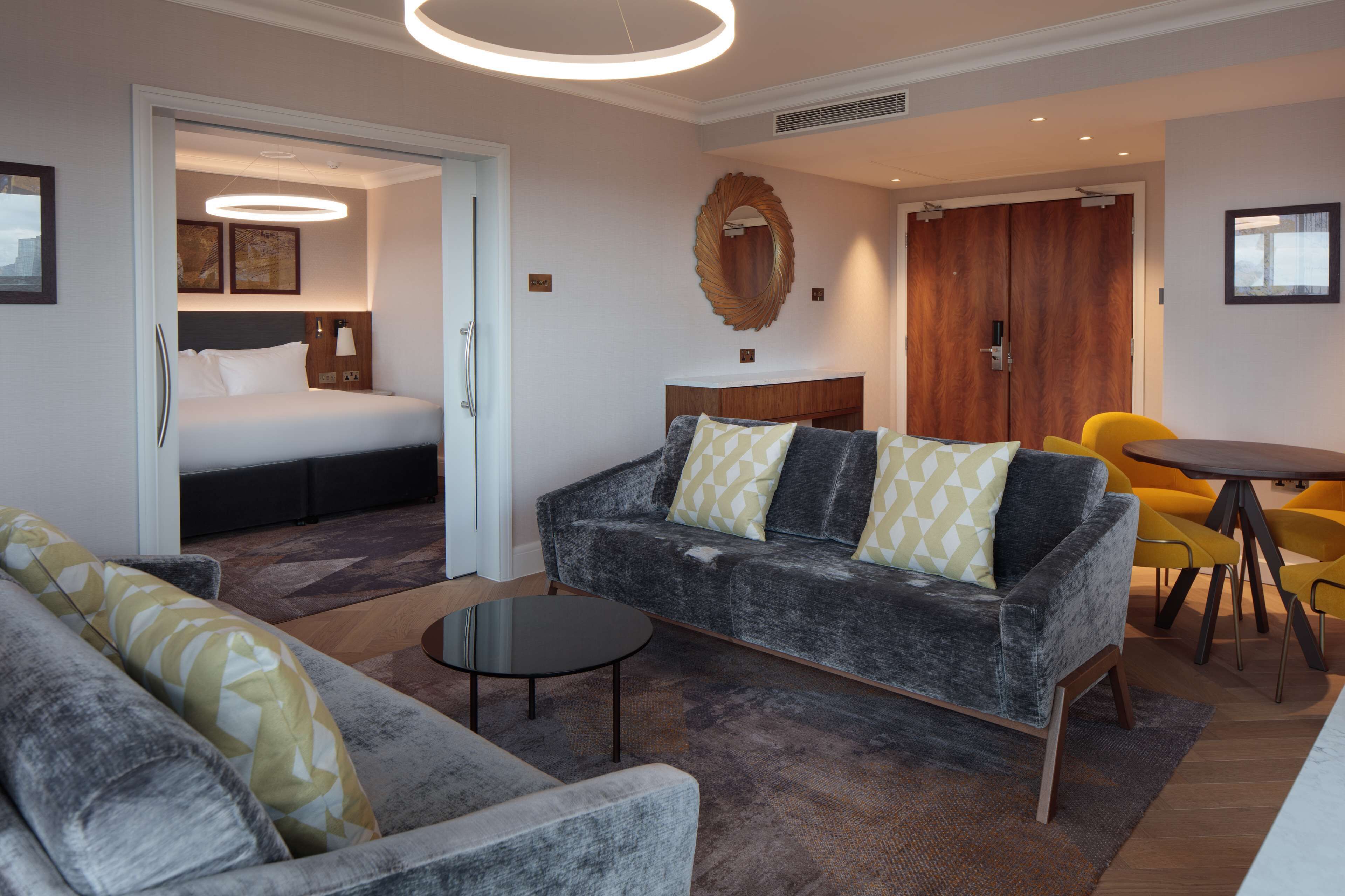 A modern hotel room with a grey sofa, a small round table, a bed with a headboard, a wooden wardrobe, and a mirror on the wall.