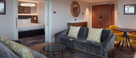 A modern hotel room with a grey sofa, a small round table, a bed with a headboard, a wooden wardrobe, and a mirror on the wall.