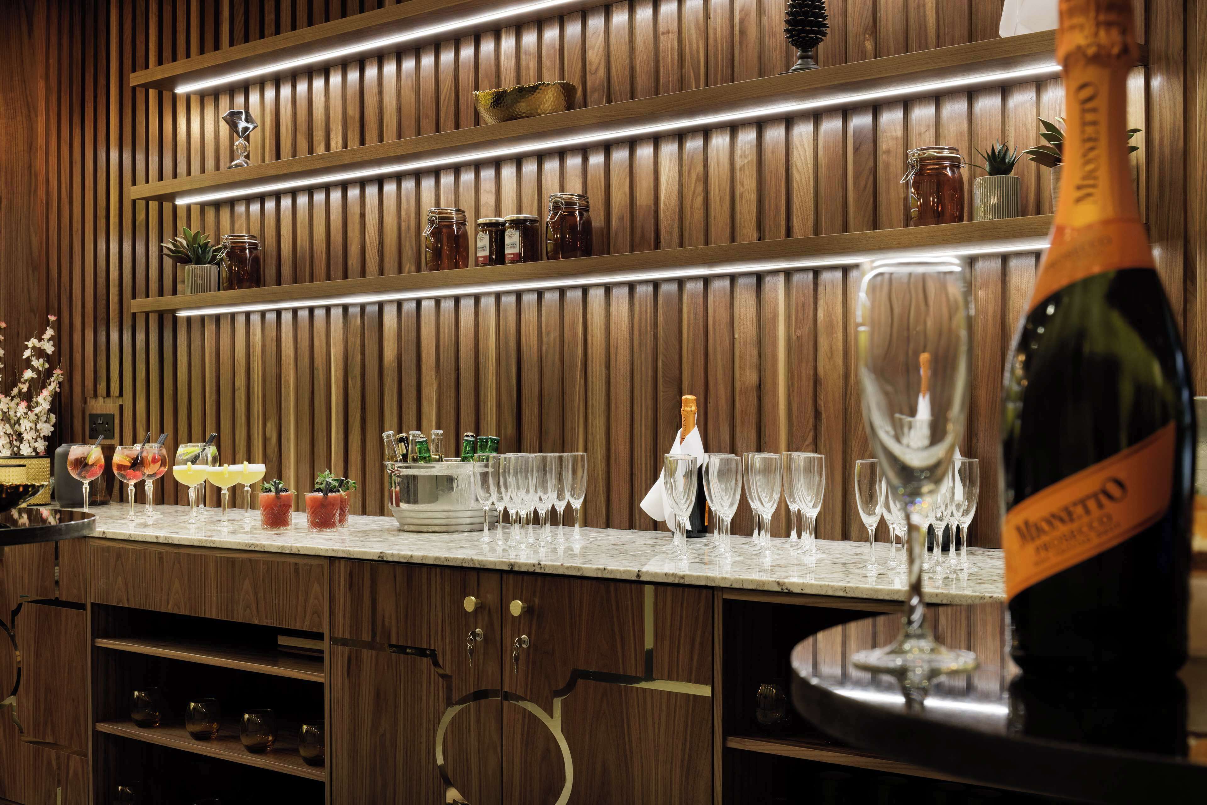 Una zona de bar con paneles de madera, estantes con frascos y una botella de champán.