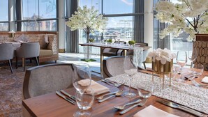 A dining area with a wooden table set for a meal, surrounded by chairs, with a view of a bridge through large windows.