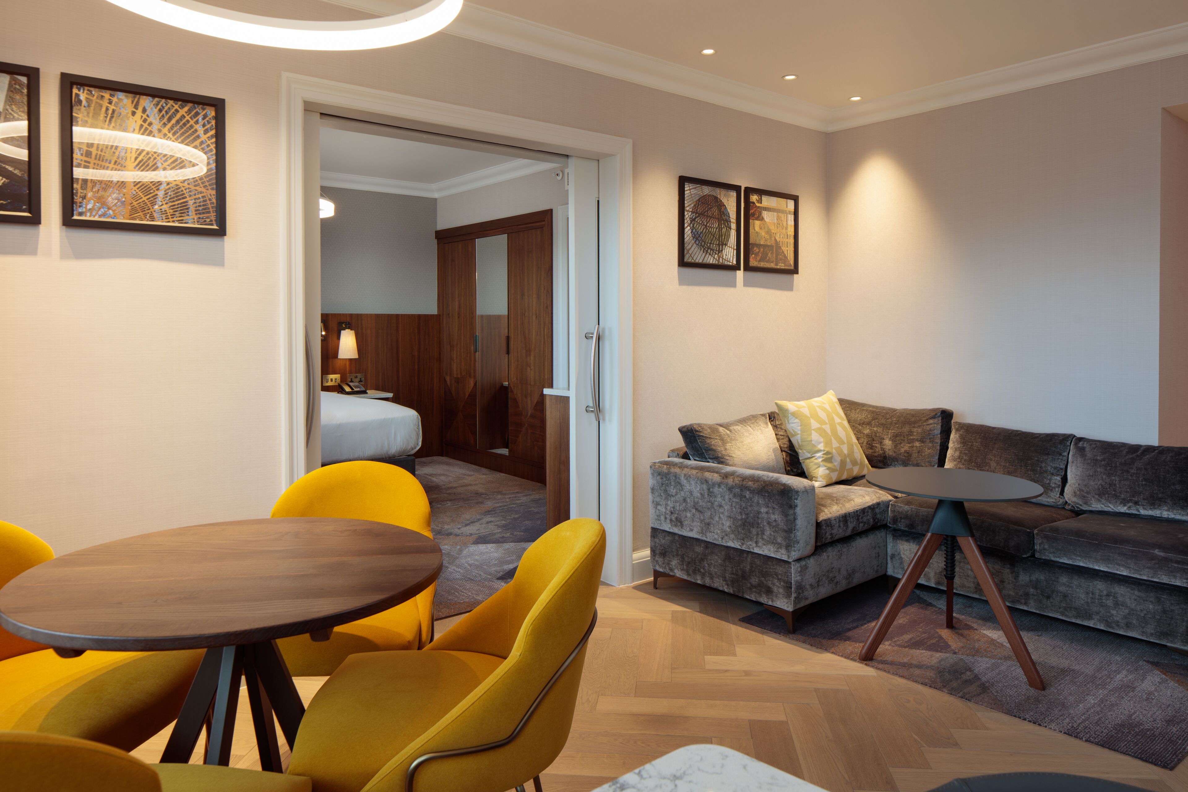 A modern hotel room with a sofa, yellow chairs, a round table, and artwork on the walls.