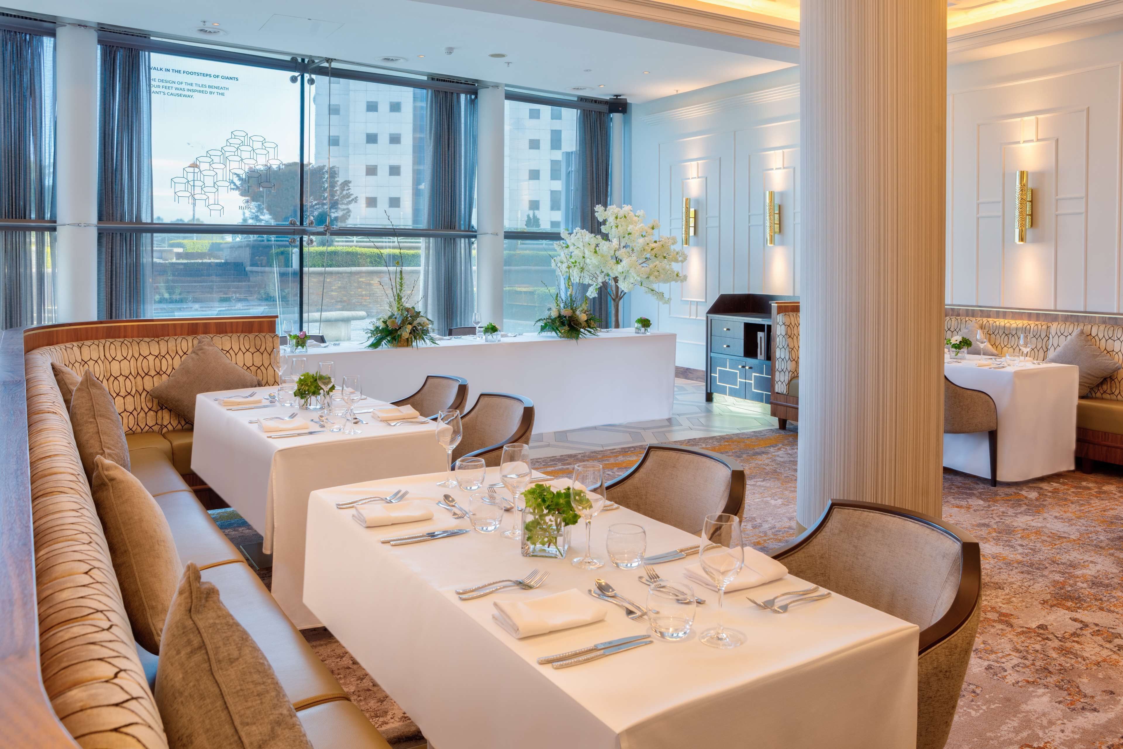 En restaurant i et hotel med dækkede borde, store vinduer og udsigt over bybilledet.