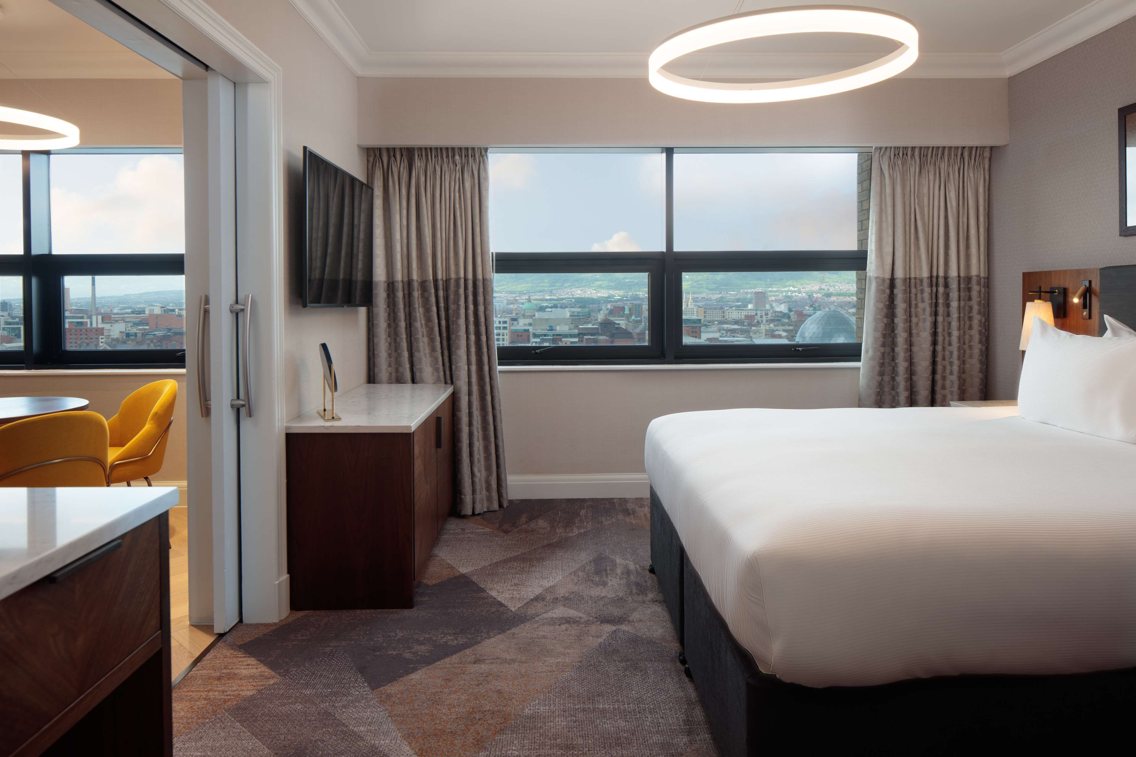 Habitación de hotel con una cama grande, un escritorio, una silla, un televisor y vistas a la ciudad a través de la ventana.