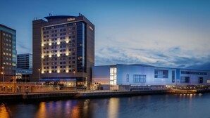 A modern hotel with a prominent Hilton sign, a waterfront location, and a contemporary building adjacent to it.