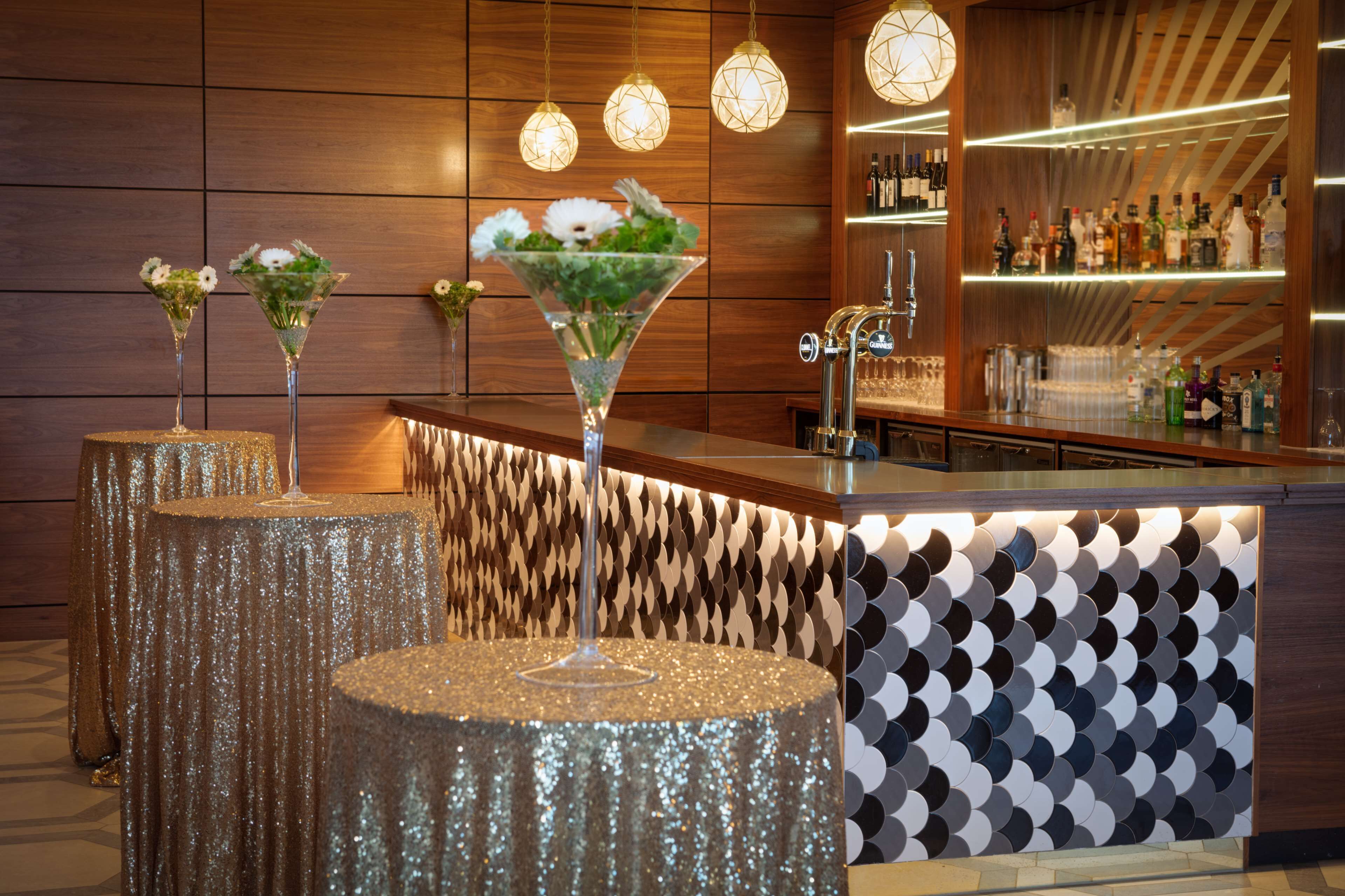 Zona de bar con pared de madera, tres lámparas colgantes y una barra con botellas y vasos.