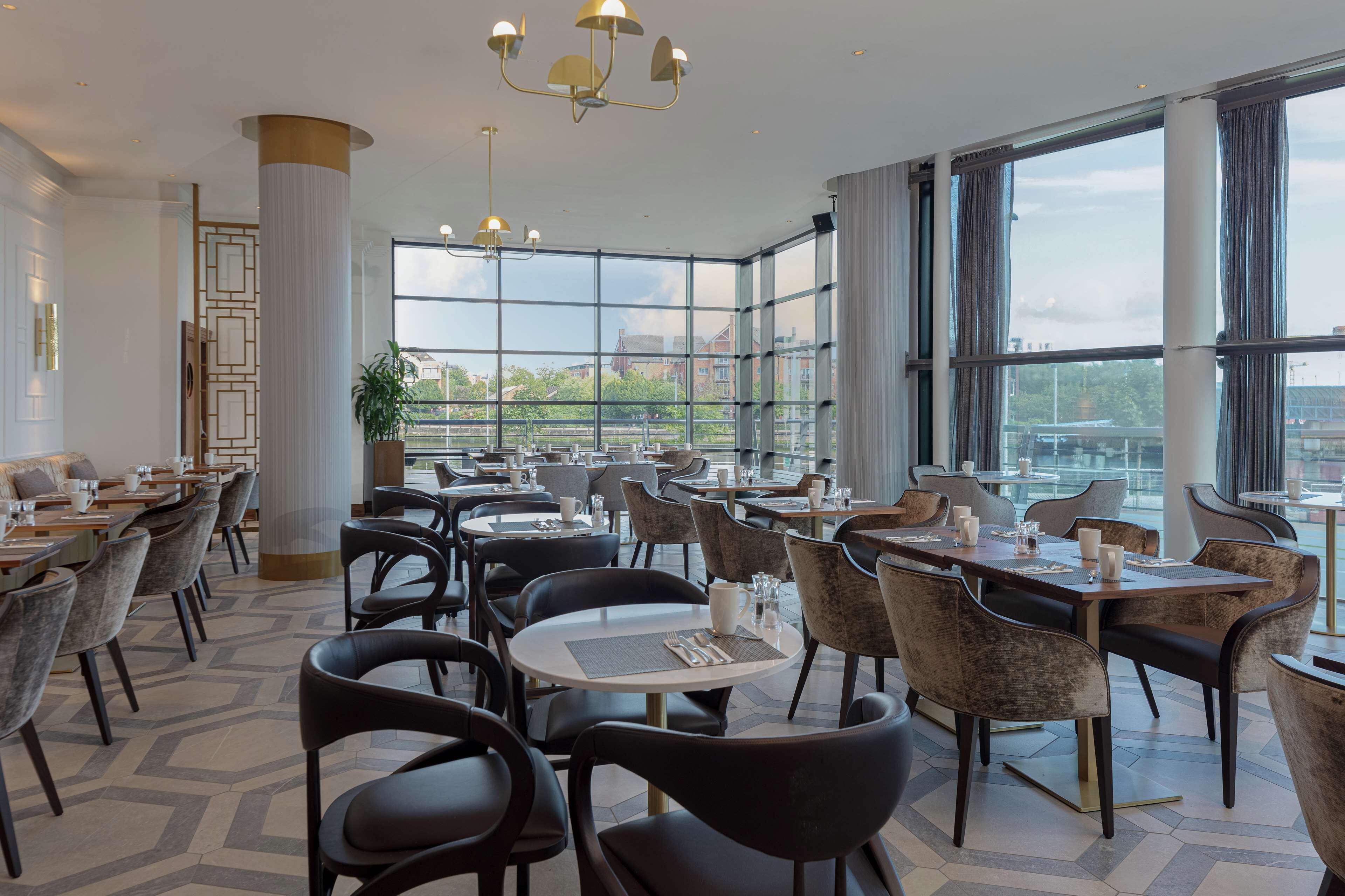 A spacious dining area with multiple tables and chairs, large windows offering a view of the cityscape, and modern lighting fixtures.
