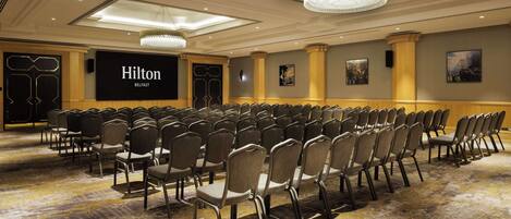 Un salón de conferencias con filas de sillas, una pantalla grande que muestra 'Hilton Belfast' y un candelabro.