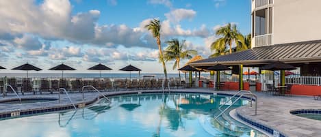 Une piscine entourée de palmiers et d’un bâtiment doté de balcons, avec des chaises longues et des parasols à proximité.