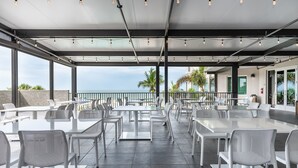 An outdoor dining area with white tables and chairs, a covered ceiling with hanging lights, and a view of the ocean. - DiamondHead Beach Resort (Fort Myers Beach)