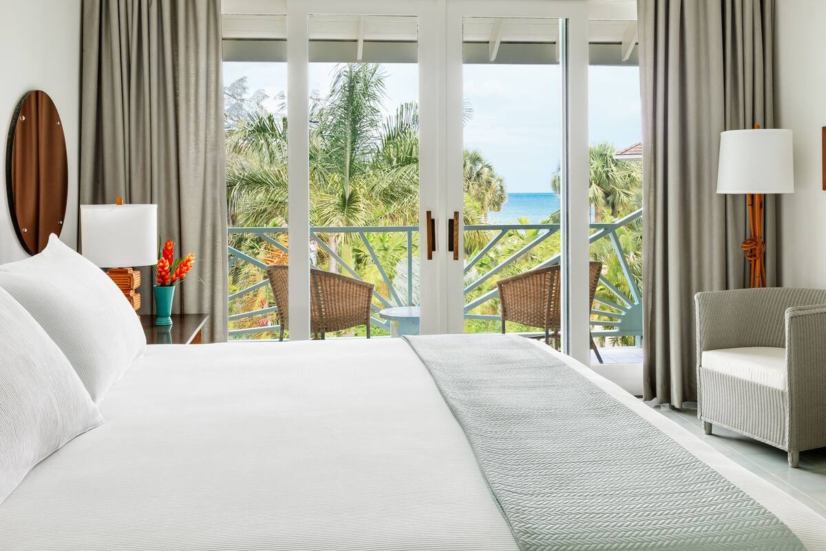 A hotel room with a large bed, a balcony with wicker chairs, and a view of the ocean and palm trees.