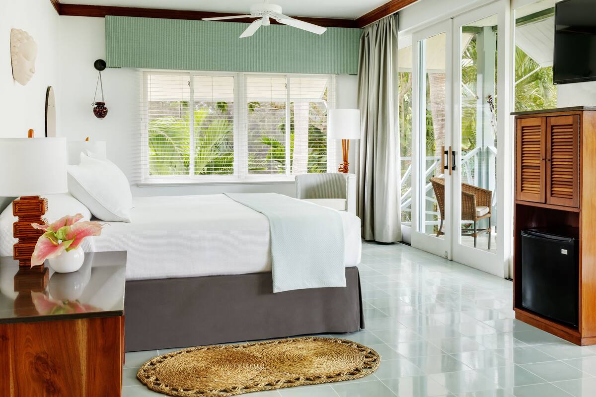 A bedroom with a bed, a desk, a lamp, a TV, and a sliding glass door leading to a balcony.
