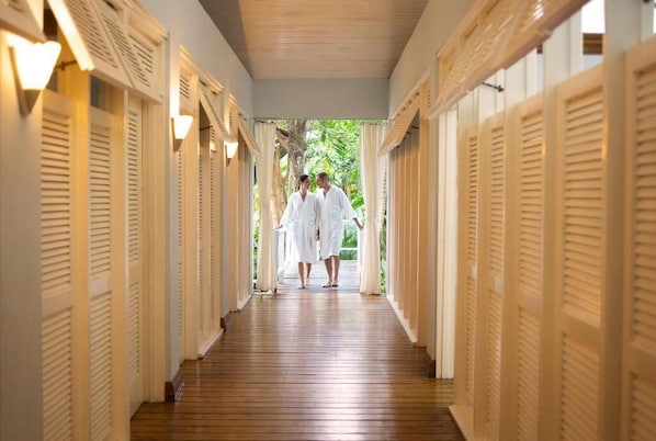 A corridor with wooden flooring and white doors with shutters, leading to an outdoor area with greenery. - Couples Negril All Inclusive (Negril)