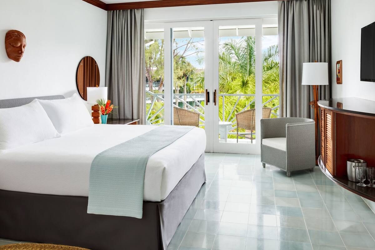 A hotel room with a bed, a seating area with a chair, a TV, and a balcony with a view of greenery.