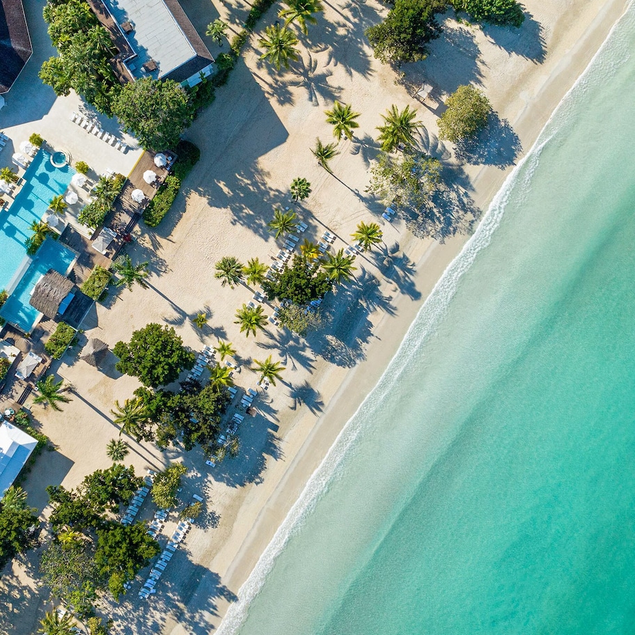 An aerial view of a beach resort with a swimming pool, sun loungers, and palm trees.