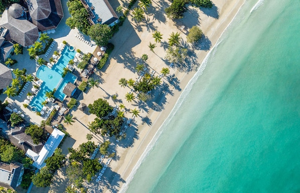 Vue aérienne d’un complexe hôtelier de bord de mer, avec une piscine, des chaises longues et des palmiers.