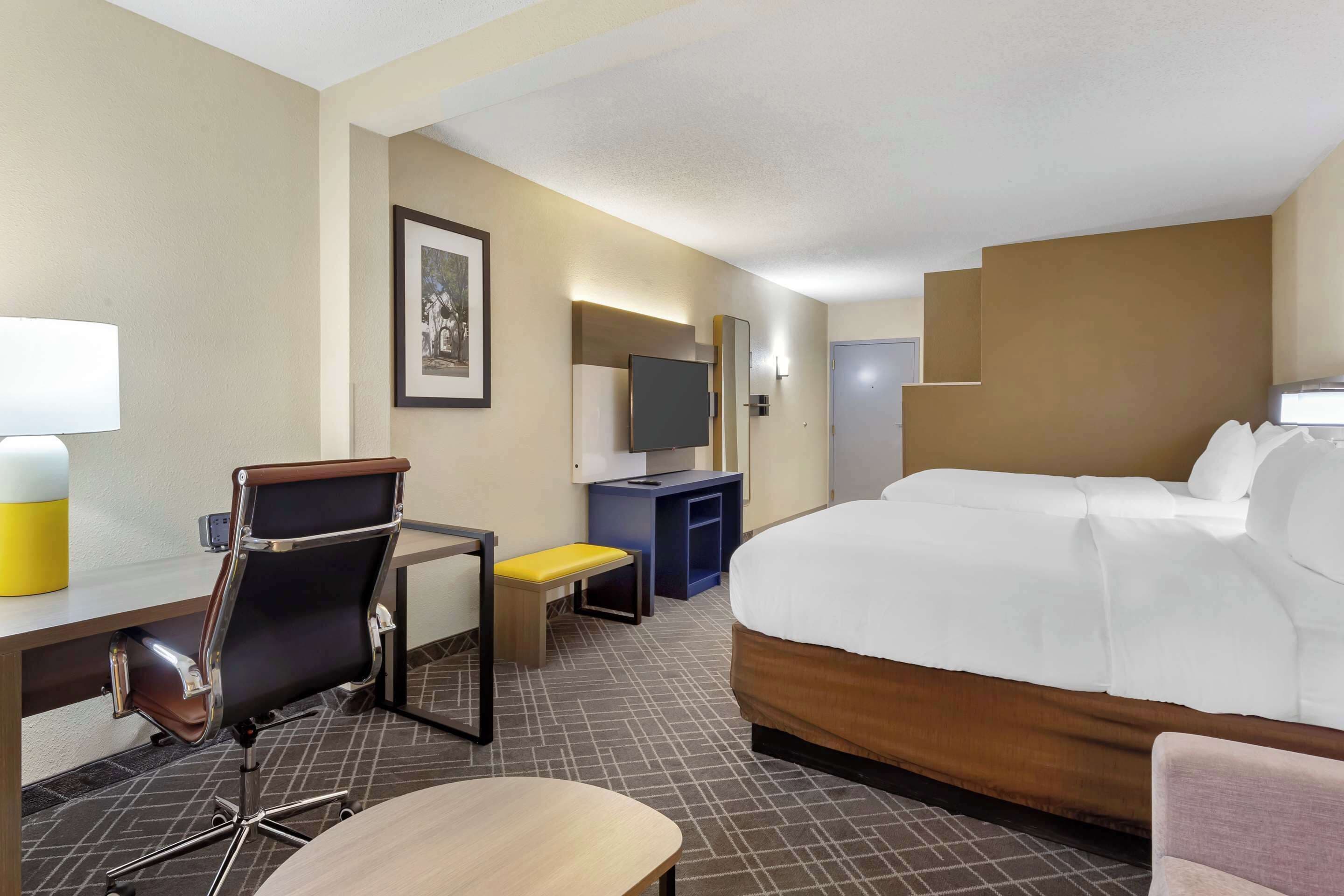 A hotel room with a large bed, a desk with a chair, a small table, a TV, and a framed picture on the wall.