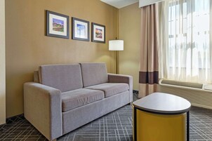 Habitación de hotel con sofá gris, una mesita redonda y una lámpara de pared. Se ven tres cuadros en la pared y una ventana con cortinas.