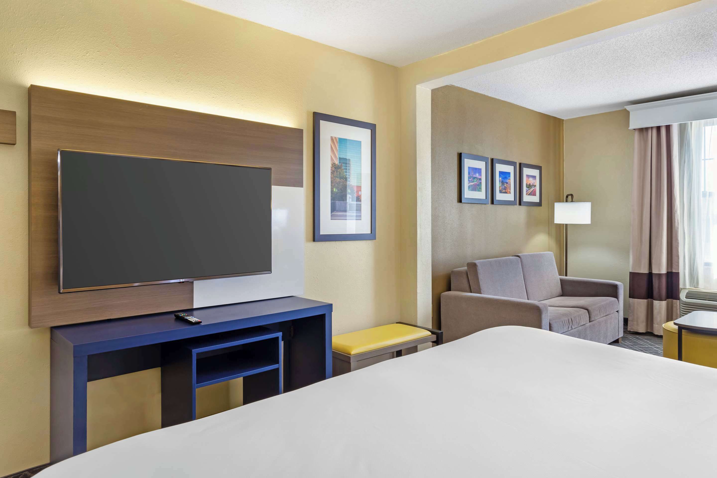 A hotel room with a large bed, a TV on a wooden stand, a grey armchair, a small table, and a lamp.