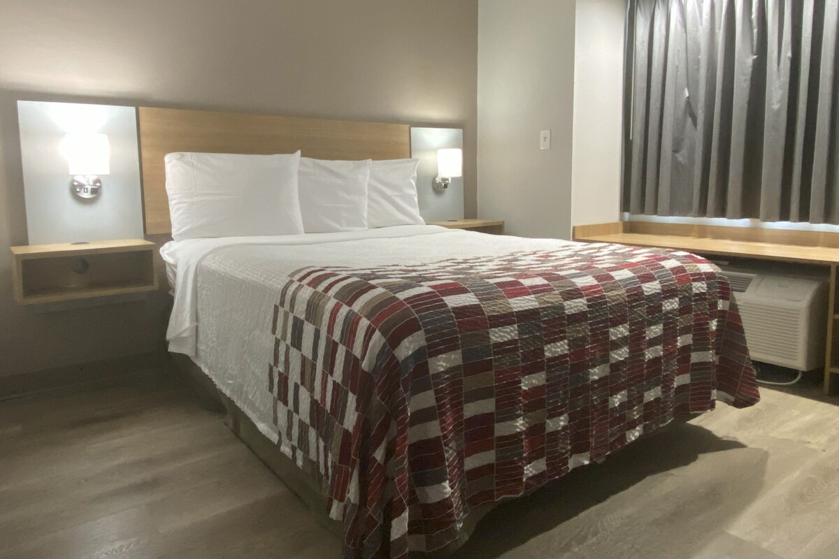 A hotel room with a bed, two bedside tables with lamps, a window with curtains, and a radiator.