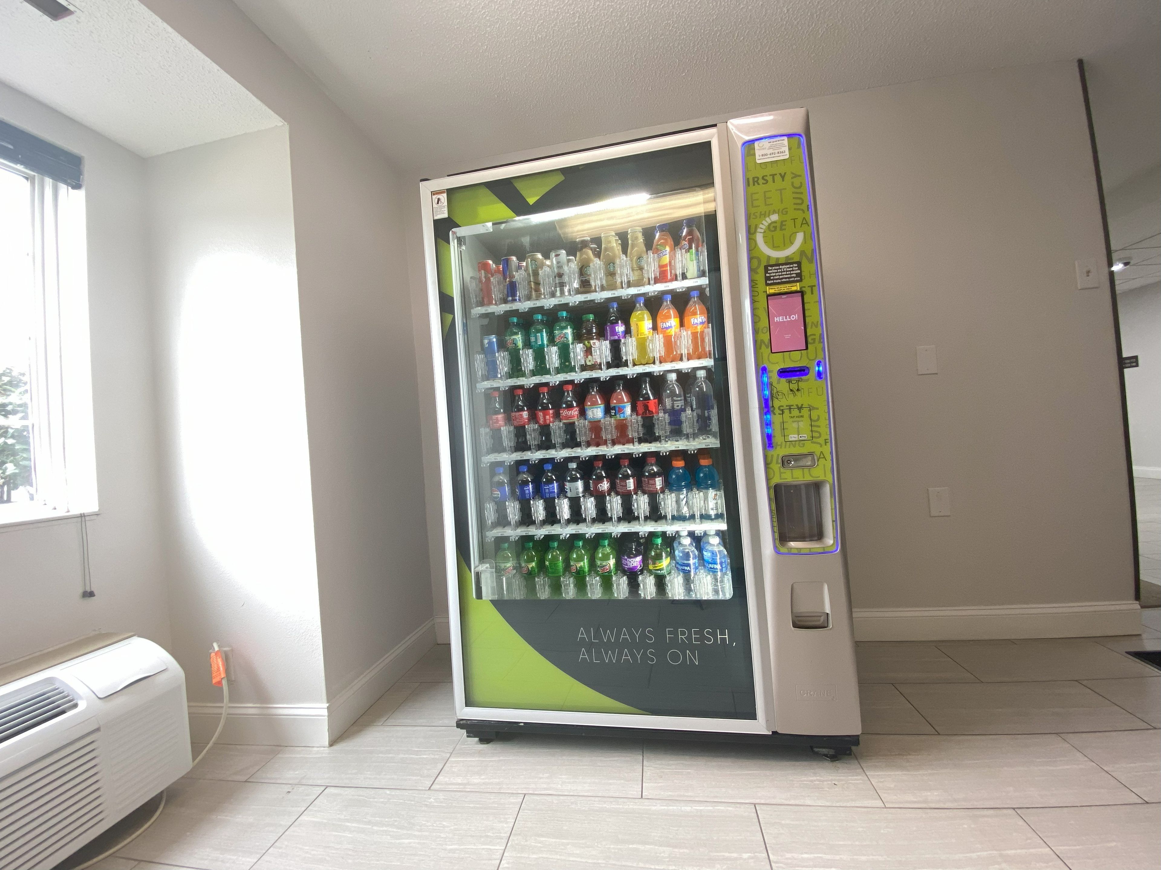 A vending machine stocked with various beverages in a room with a window and a wall-mounted air conditioner.