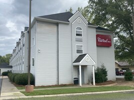 Um hotel com uma placa vermelha que diz "Red Roof Inn". Apresenta um edifício grande e de vários andares com uma entrada imponente e um pequeno alpendre.