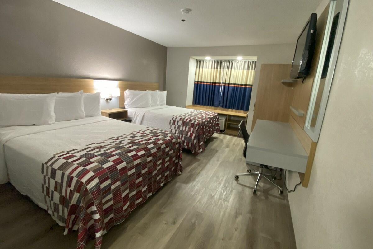 A hotel room with two beds, a desk, a chair, a TV, and a window with curtains.