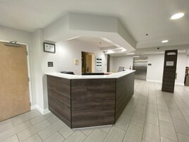 Zona de recepción con un mostrador de madera oscura, una puerta y vista a un pasillo con un refrigerador y otra puerta.