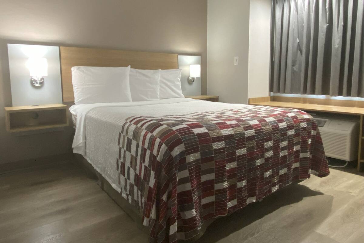 A hotel room with a bed, two bedside tables with lamps, a window with curtains, and a radiator.