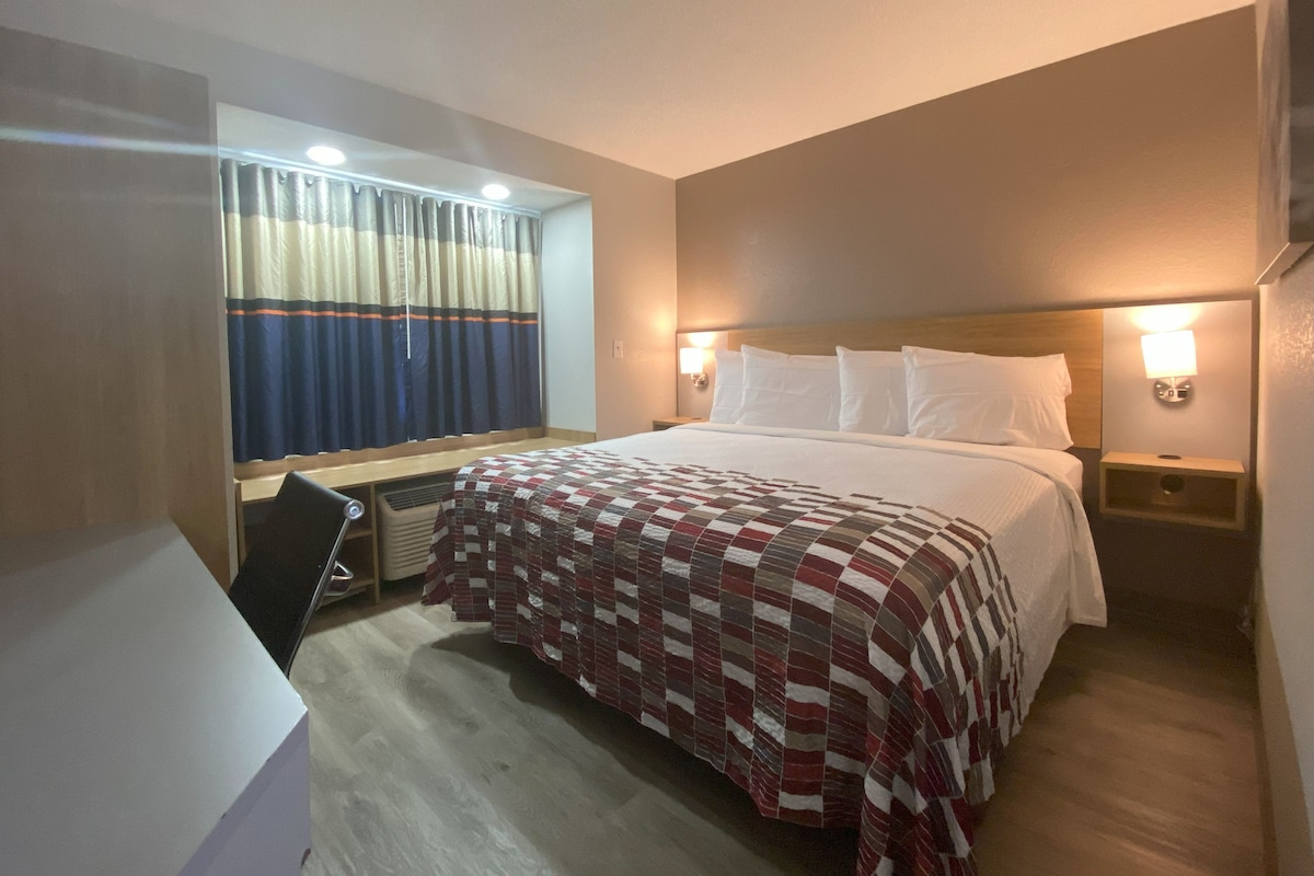 A hotel room with a bed, a desk, a chair, a window with curtains, and a nightstand with a lamp.