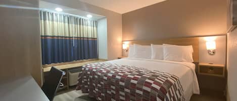 Habitación de hotel con cama, escritorio, silla, ventana con cortinas y mesita de noche con lámpara.