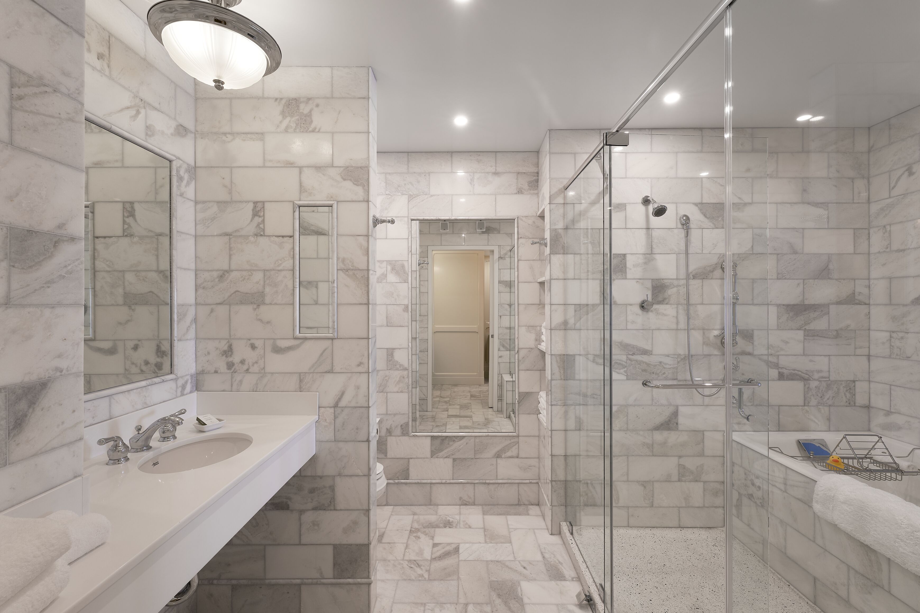 Un baño moderno con ducha revestida de mármol, zona de ducha con mampara de vidrio y tocador con lavabo y espejo.