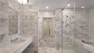 Un baño moderno con ducha revestida de mármol, zona de ducha con mampara de vidrio y tocador con lavabo y espejo.